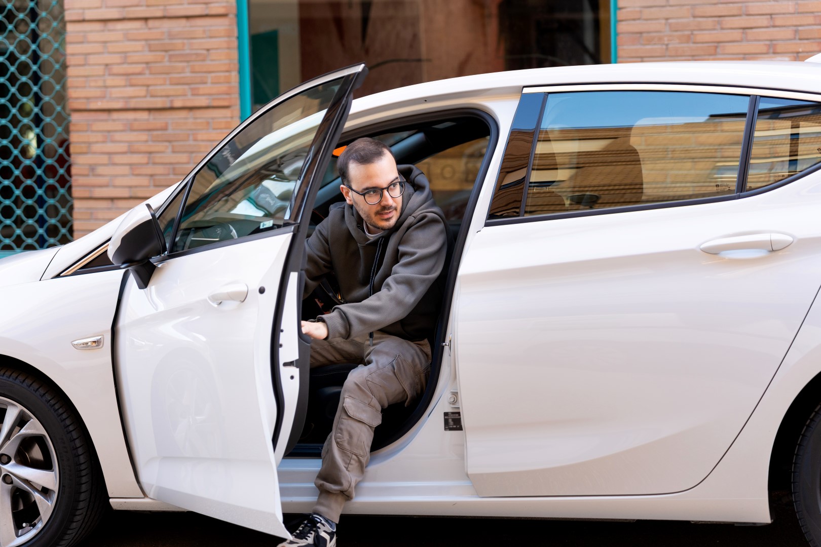 Young man wearing hoodie and glasses, exiting modern white car on a downtown street, starting a workday
